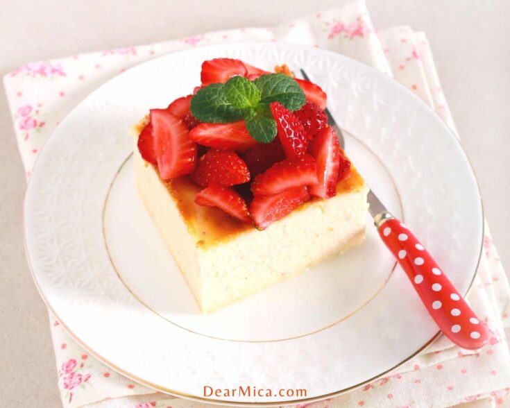 Top view of a slice of no crust cheesecake topped with fresh strawberries and mint, served on a white plate with golden rims and a red white polka dot fork.