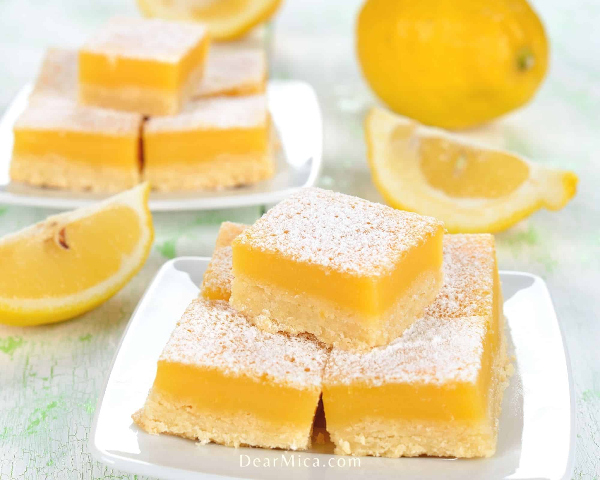 Side view of homemade keto lemon bars sprinkled with powder keto-friendly sugar served stacked up on a white plate on table top with lemons and lemon wedges propped on the background.