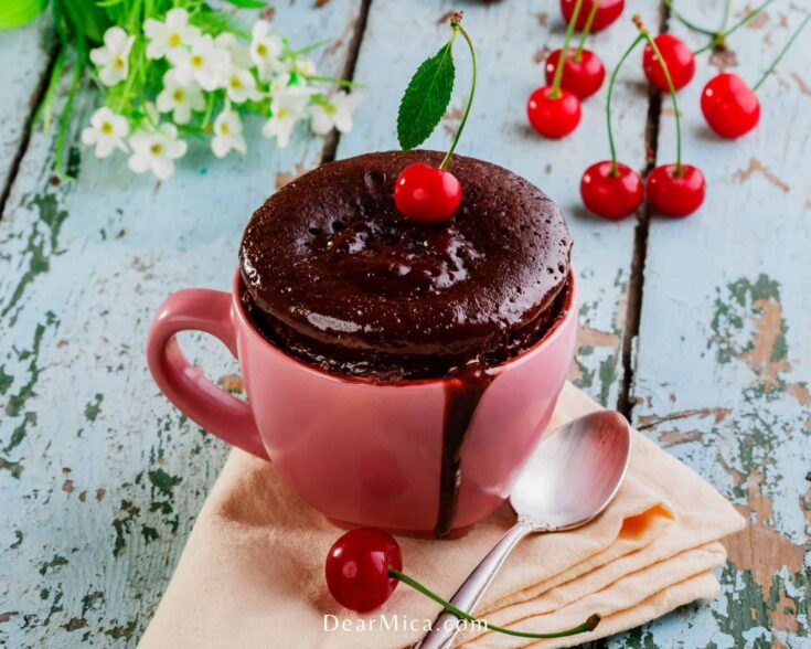 A Keto Chocolate Mug Cake in a pink mug garnished with 1 cherry on a wooden blue table.