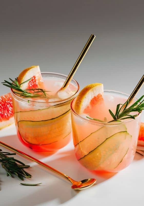 Side view of two low carb Grapefruit Gin Fizz Cocktail With Cucumber garnished with cucumber and rosemary on a white background with grapefruit wedges.