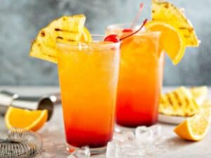 Side view of Low Carb Tequila Drinks. Two tall glasses of tequila sunrise cocktail garnished with grilled pineapple slices and cherries.