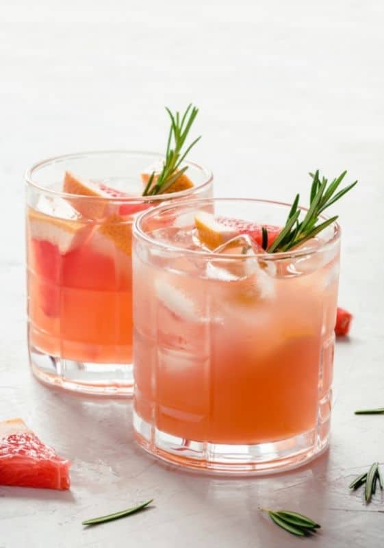 Side view of two glasses of low carb grapefruit ginger fizz cocktail garnisheed with grapefruit slices and sprig of rosemary on a gray background decorated with grapefruit wedges.