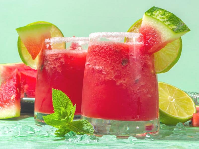 Two glasses of Keto Jalapeño Watermelon Margarita garnished with limes and watermelon on a green background.