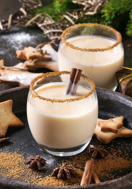 Side view of two glasses of Low Carb Eggnog Cocktail with a cinnamon stick on a holiday decorated table.