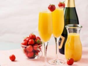 Two low carb mimosa garnished with strawberries with bottle of champagne and orange juice on the side.